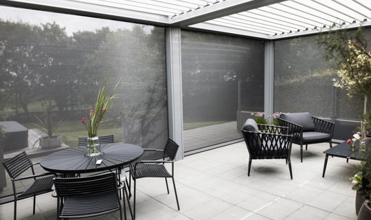 Moderna pergola z vrtljivimi lamelami in stranskimi screen roloji, opremljena z vrtnim pohištvom na tlakovanem dvorišču, obdana z zelenjem.
