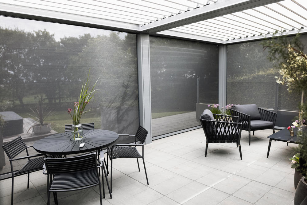 Moderna pergola z vrtljivimi lamelami in stranskimi screen roloji, opremljena z vrtnim pohištvom na tlakovanem dvorišču, obdana z zelenjem.